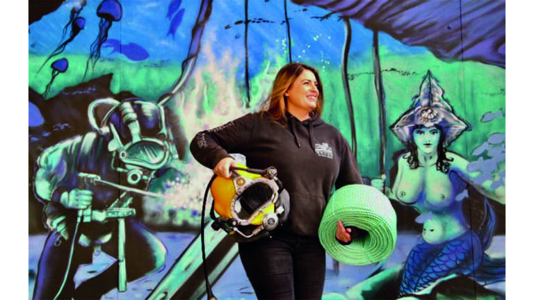 Une plongeuse souriante tient un casque de plongée devant une fresque marine colorée, symbolisant l'aventure sous-marine.