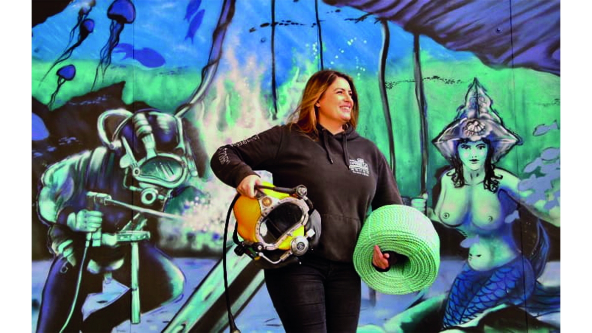 Une plongeuse souriante tient un casque de plongée devant une fresque marine colorée, symbolisant l'aventure sous-marine.