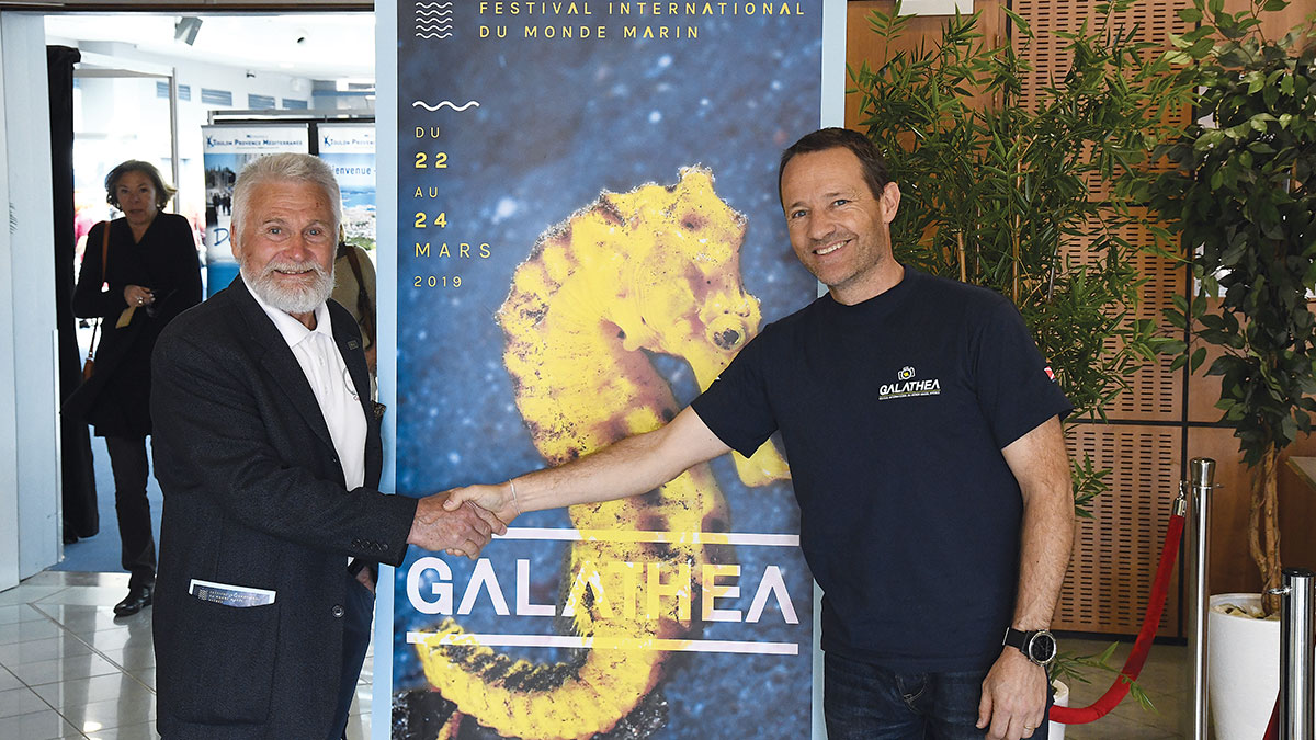 Deux hommes se serrent la main devant une affiche colorée du Festival Galathéa, célébrant la passion pour le monde marin.