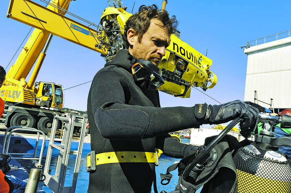 Un plongeur prépare son équipement sous le regard d'un submersible jaune en arrière-plan.