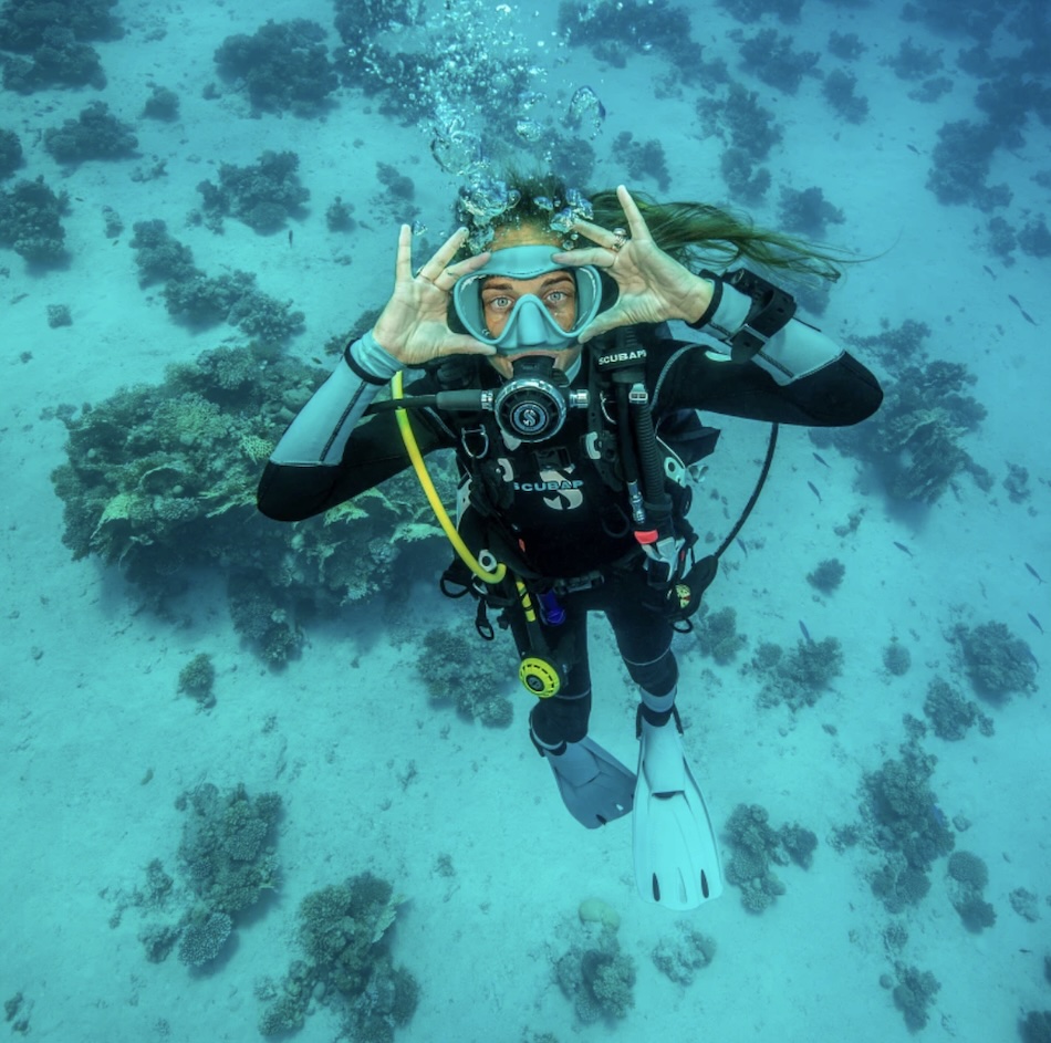 Plongeuse souriante explorant les fonds marins, entourée de coraux et de poissons colorés.